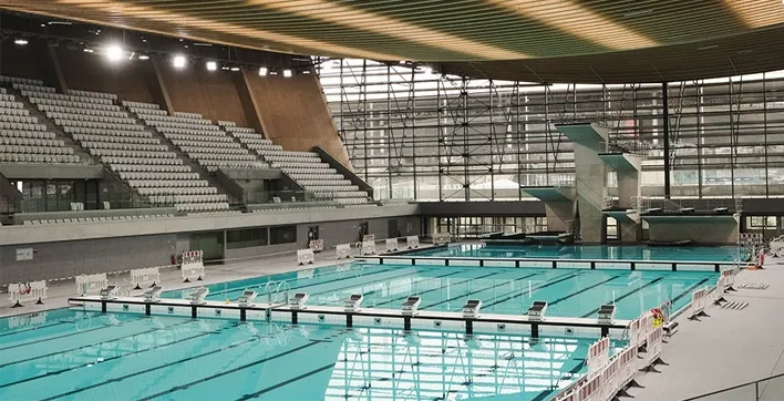 Photo du Centre Aquatique Olympique de Saint-Denis, une piscine moderne et spacieuse avec un grand bassin et un plongeoir à plusieurs hauteurs, baignée de lumière. Elle est reliée au Stade de France par une passerelle visible en arrière-plan.